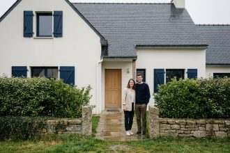 Couple devant leur maison neuve en Bretagne avec toiture en ardoise et volets bleus marine