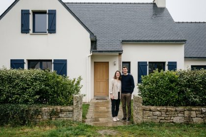Couple devant leur maison neuve en Bretagne avec toiture en ardoise et volets bleus marine